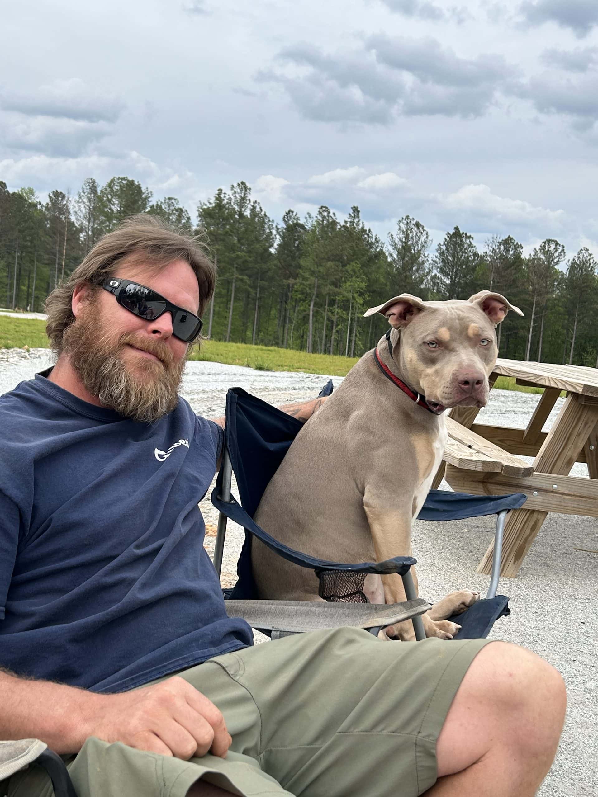 A man with sunglasses and a beard relaxes in a camp chair next to a gray and tan dog sitting upright in its own chair, both enjoying the outdoors near a picnic table and gravel lot. Pine trees and a cloudy sky frame the backdrop, capturing a laid-back RV travel moment with a furry companion.