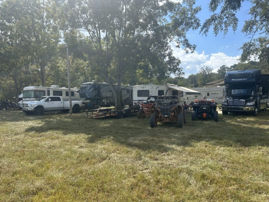 Several RVs and campers, including a black Pasadena motorhome and a fifth-wheel trailer, are parked under tall trees on a grassy field, with a white truck and several muddy ATVs in front. This off-grid setup showcases a group boondocking site surrounded by nature, ideal for RV travel and outdoor recreation.