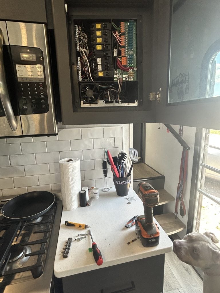 An RV kitchen counter covered in tools and parts sits beneath an open electrical panel mounted in a cabinet, showing exposed wiring and circuit breakers. A dog looks up curiously at the workspace, adding a bit of personality to this behind-the-scenes moment of RV repairs.