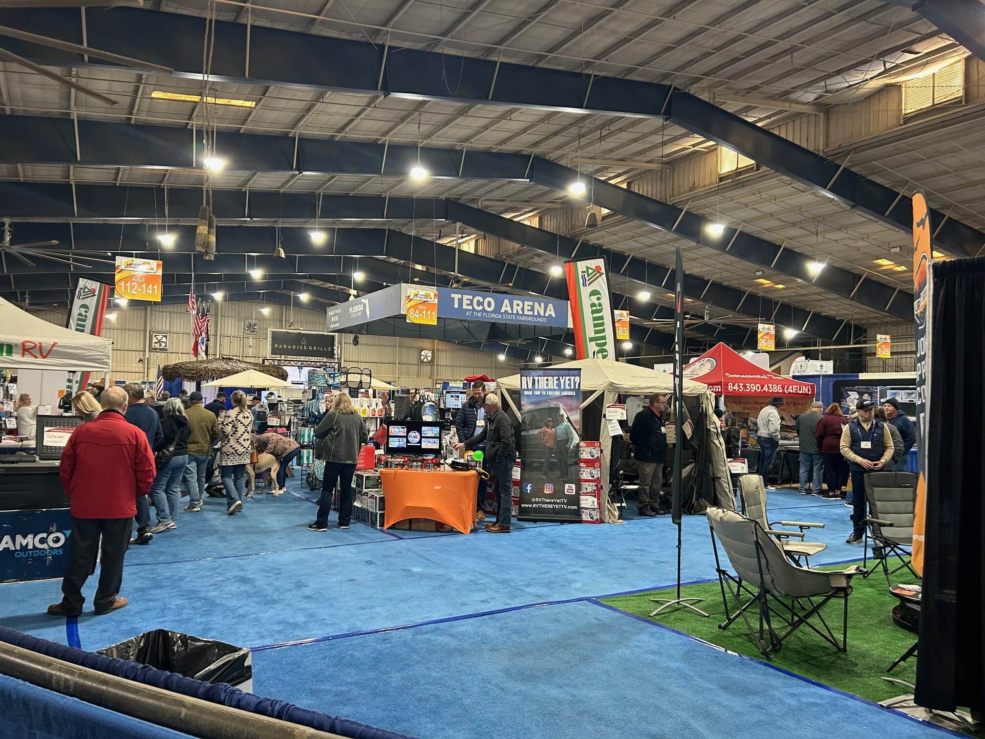 Indoor RV show at TECO Arena with several vendor booths, including RV There Yet? and Camco Outdoors, surrounded by attendees browsing products and displays. The floor is covered in blue carpet, and camping chairs, flags, and banners add to the event atmosphere.