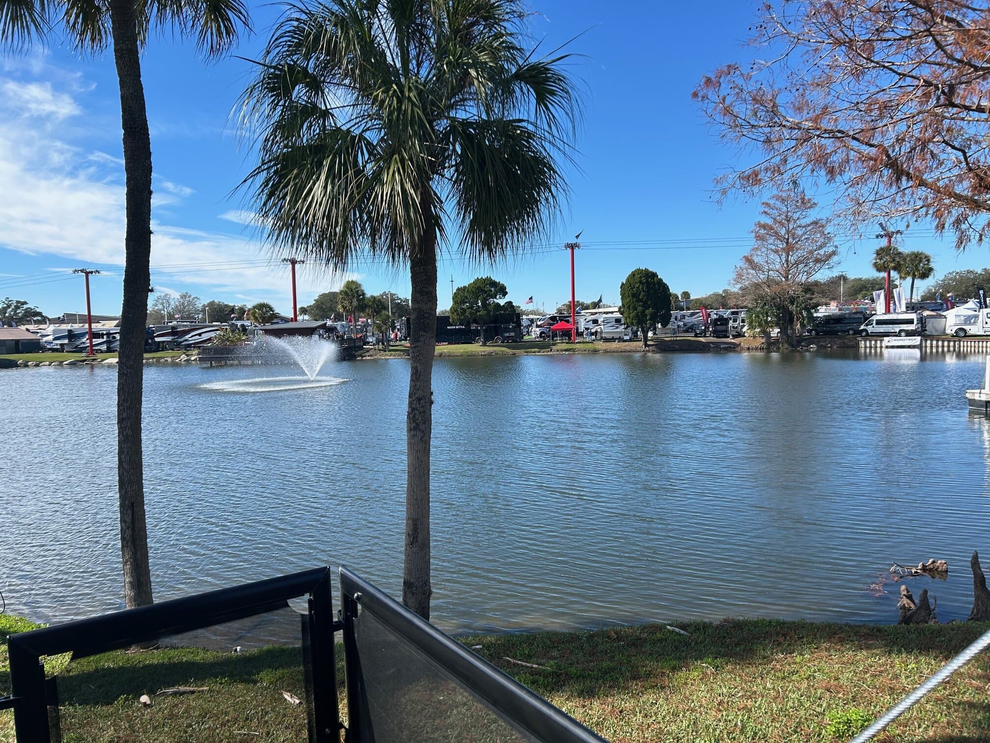 Scenic view of a small lake with a central fountain spraying water, surrounded by palm trees and grassy edges. Across the water, an RV show is visible with rows of vehicles, red flags, and vendor tents under a clear blue sky.