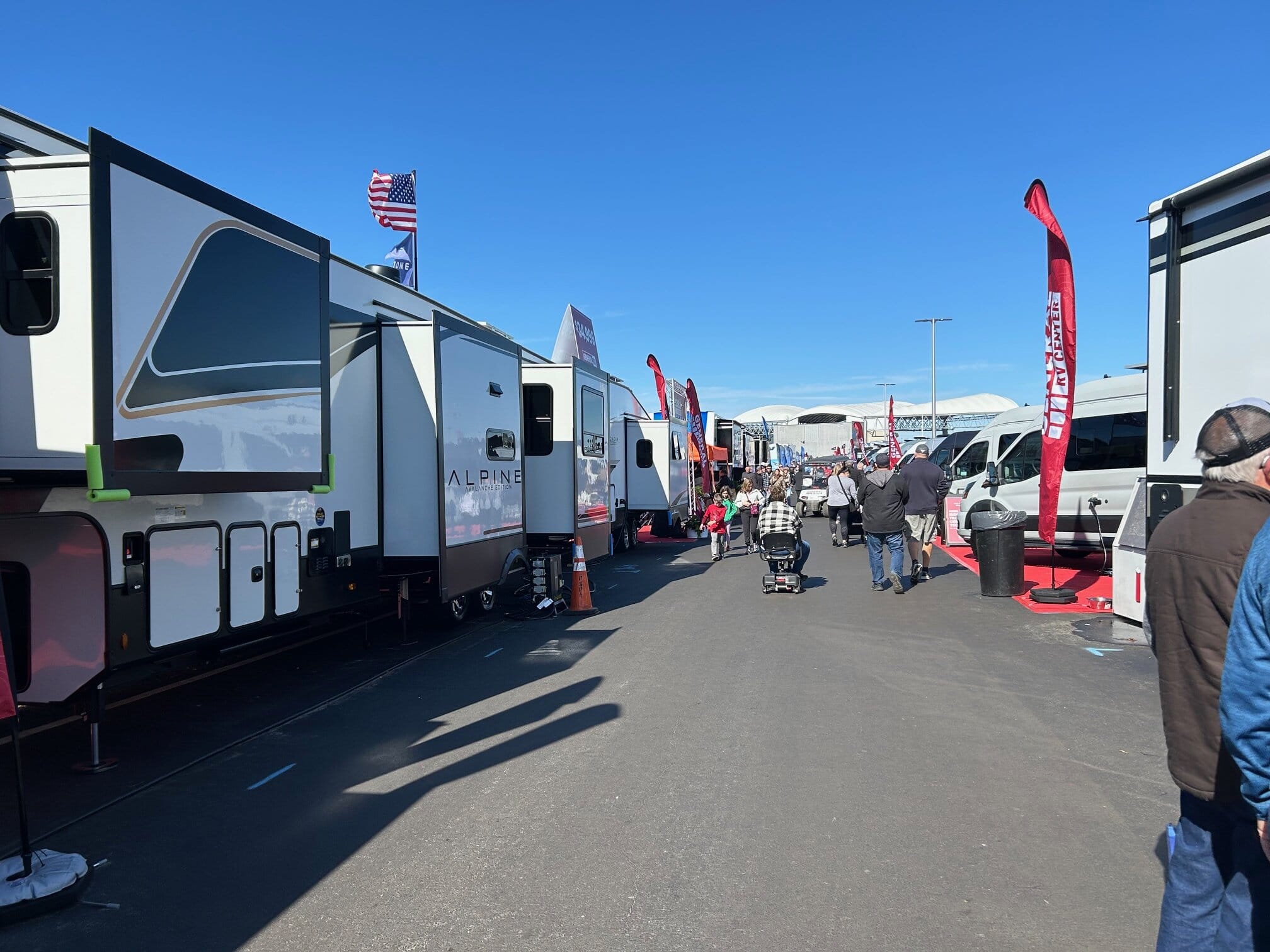 Outdoor RV show featuring rows of large RVs and trailers, including an Alpine model, with visitors walking and browsing under a clear blue sky. Red flags, vendor signs, and American flags add color to the busy asphalt pathway between displays.