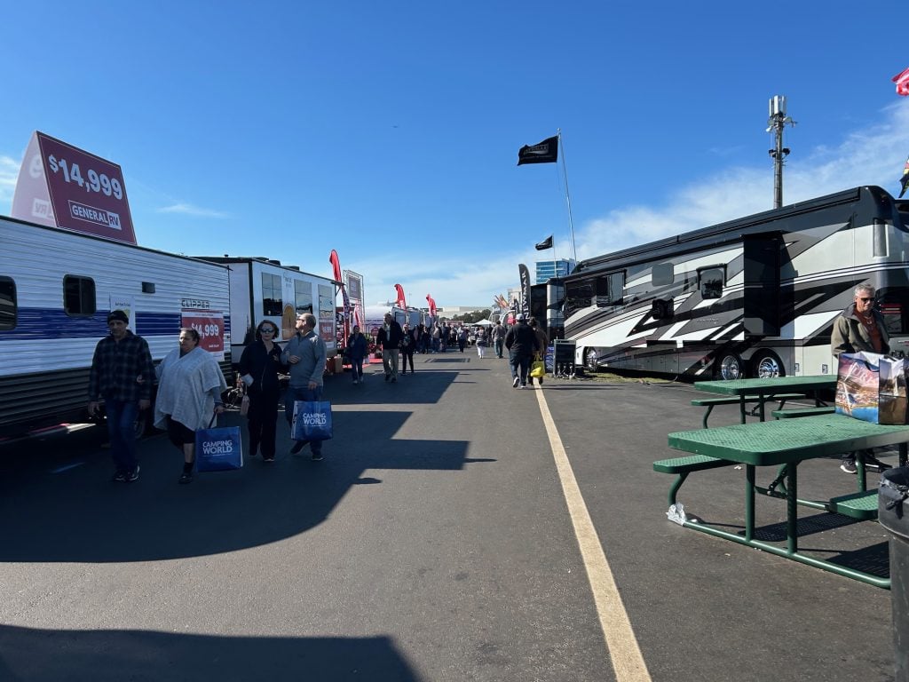 Crowds walk through an outdoor RV show lined with various trailers and motorhomes, including a Clipper trailer priced at $14,999 and a sleek black-and-silver luxury RV. Shoppers carry blue Camping World bags, and green picnic tables sit nearby under a clear blue sky.