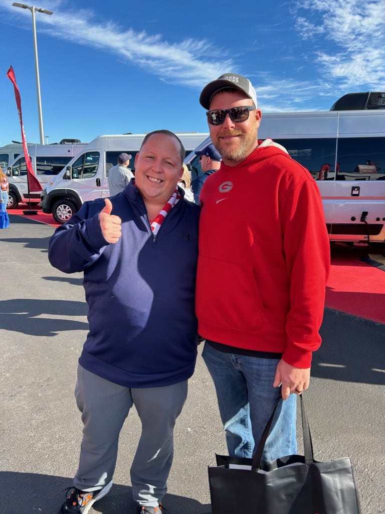 Two smiling people pose outdoors at an RV show under a bright blue sky, with vans and red carpet visible in the background. One wears a red hoodie and sunglasses, while the other gives a thumbs up and wears a navy jacket with a red-and-white lanyard.