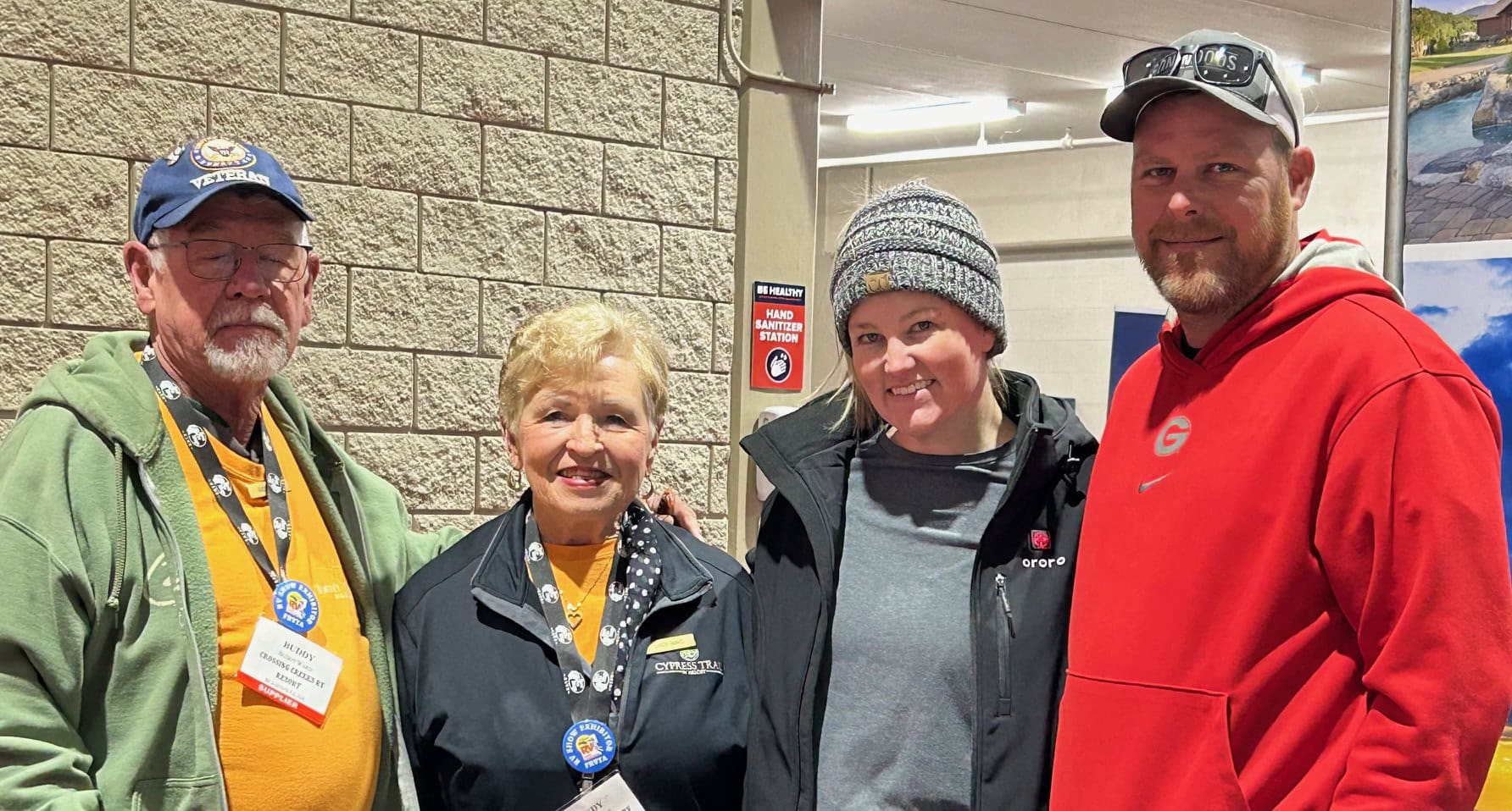 Four people pose together indoors at an RV event, smiling in front of a beige brick wall with a hand sanitizer station sign. The two on the left wear name tags and Cypress Trail Resort badges, while the two on the right are dressed in casual winter clothing, including a red hoodie and a knit hat.