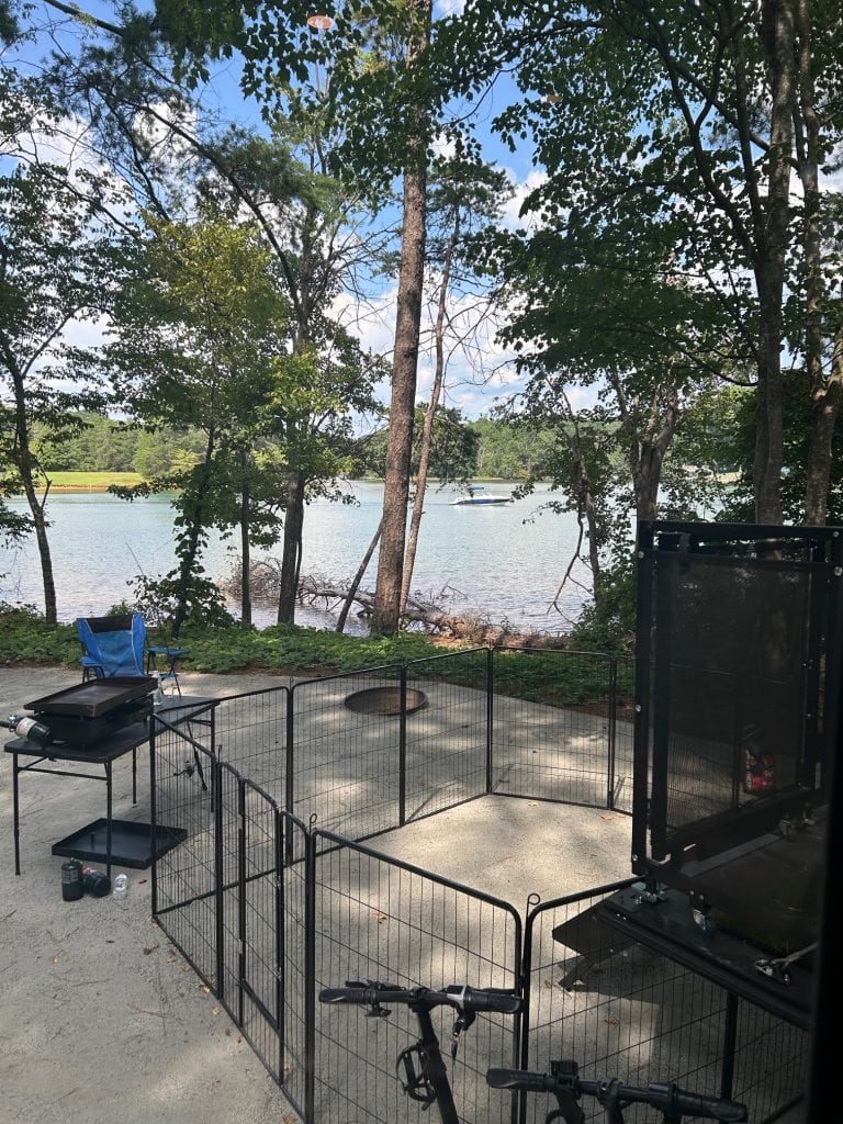 View from a shaded lakeside campsite featuring a fenced-in patio area, a blackstone grill setup, and a blue camping chair overlooking the water. In the background, a small boat floats on the lake, partially visible through tall trees under a bright, partly cloudy sky.