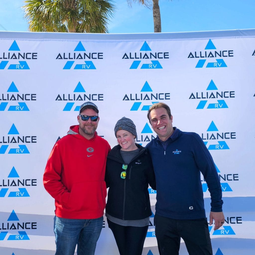 Three people smile in front of a branded Alliance RV backdrop at an outdoor event, with palm trees and blue sky in the background. The man on the left wears a red hoodie and sunglasses, the woman in the center wears a knit hat and black jacket, and the man on the right wears a navy Alliance RV pullover.