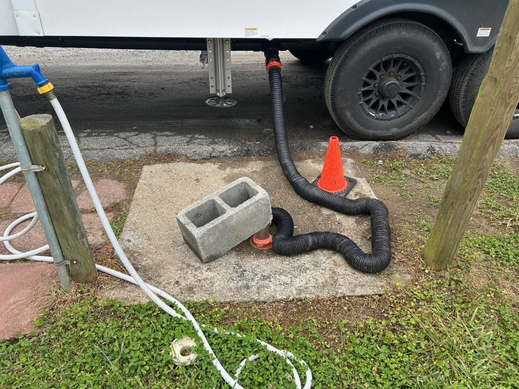 An RV sewer hose setup at a campsite, with a black flexible drain hose connected from the RV to the ground sewer inlet. A concrete cinder block and orange traffic cone are used to help secure the hose in place, while a white freshwater hose is seen on the left near a blue spigot.