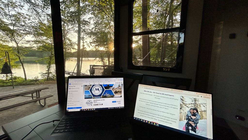 A cozy RV workspace setup with an open laptop and a Xebec Soloscreen on a table near a large window, overlooking a serene lakeside campsite surrounded by trees at sunset. One screen displays a Facebook page for "Middletons of the Move" and the other shows a blog introducing RV travel experts, with a photo of a couple in snow gear. The peaceful natural view contrasts with the digital work setup inside.