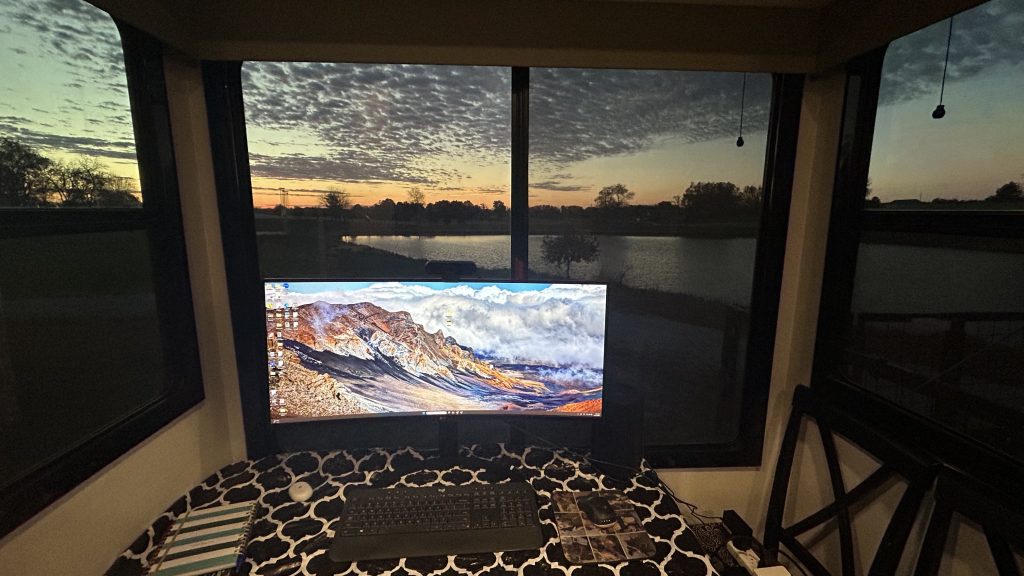 An RV workspace with a large curved monitor on a patterned black and white tablecloth, framed by panoramic windows revealing a peaceful lake view at sunset. The desktop displays a vivid mountain landscape, and a keyboard, mouse, notebook, and coasters are neatly arranged on the table. The glowing sky and reflection on the water create a serene backdrop for remote work.
