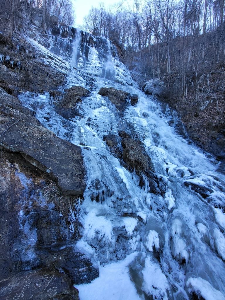 A dramatic winter scene at Amicalola Falls in North Georgia shows the cascading waterfall partially frozen, with sheets of ice and icicles clinging to the rocky slope. Bare trees line the steep hillside, and mist rises from the flowing sections of water that remain unfrozen.