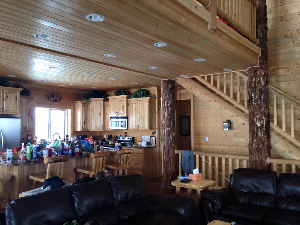 Interior of a rustic log cabin with knotty pine walls, ceiling, and cabinetry, featuring a cozy open-concept kitchen and living area. The kitchen island is cluttered with snacks and drinks, and the space includes leather sofas, exposed wooden beams, and a staircase leading to a loft.