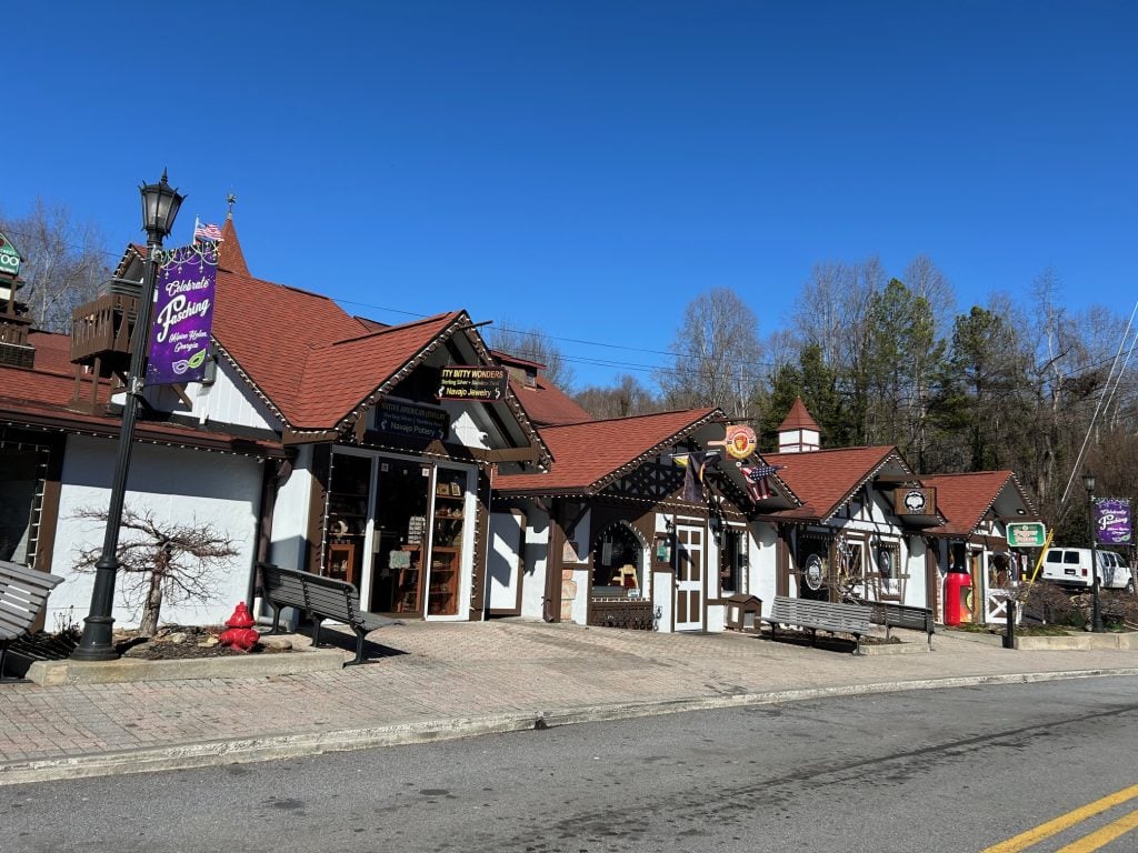 Row of charming Bavarian-style shops in downtown Helen, Georgia, featuring timber-framed facades, red roofs, and colorful signs for local businesses including Native American crafts and specialty boutiques. The cobblestone sidewalk, decorative lamp posts, and clear blue sky enhance the alpine village atmosphere that Helen is known for.