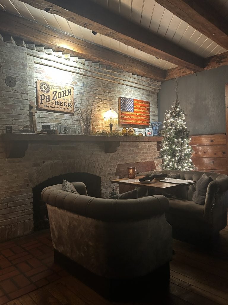 A cozy indoor dining nook at The Brewery Lodge in Michigan City, Indiana, featuring plush velvet seating, a brick fireplace, and warm ambient lighting. A lit Christmas tree and rustic decor enhance the charm of this Harvest Host stop, blending comfort with seasonal ambiance.