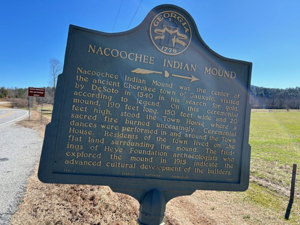 Historic roadside marker for the Nacoochee Indian Mound near Helen, Georgia, detailing its significance as a ceremonial site for the ancient Cherokee town of Gauxule. The sign highlights the mound's dimensions, cultural importance, and early exploration by archaeologists, set against a backdrop of open fields and distant wooded hills.