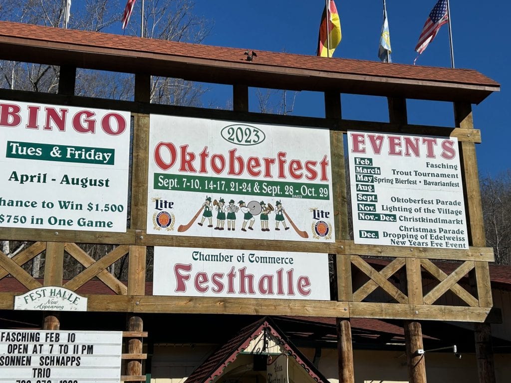 A large wooden signboard in Helen, Georgia advertises the 2023 Oktoberfest at the Festhalle, listing event dates from September 7 to October 29 and featuring playful Bavarian illustrations. Surrounding panels promote additional town events and seasonal festivities, reinforcing Helen’s German alpine village theme.