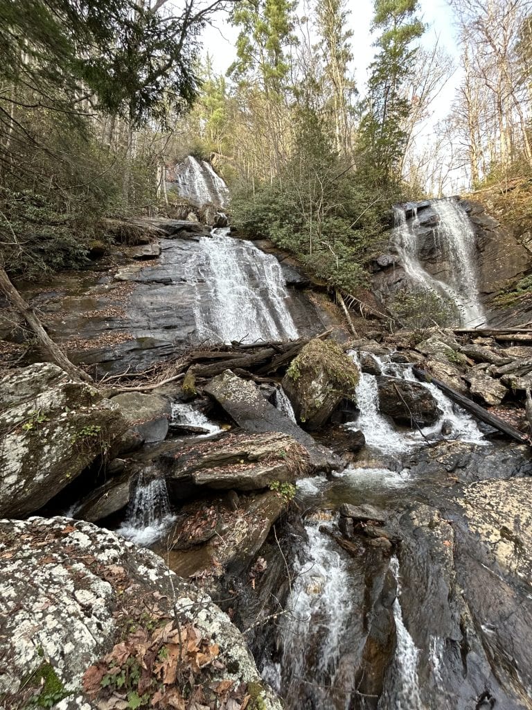Two cascading streams of water tumble down the rocky slope of Anna Ruby Falls in North Georgia, surrounded by dense evergreens and fallen leaves. The twin falls split at the top and flow side by side over smooth stone, creating a scenic, tranquil setting in the Chattahoochee National Forest.