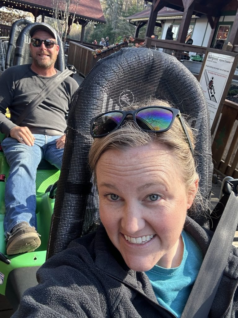 Two smiling riders are seated in individual alpine coaster cars, ready for a downhill adventure at the Georgia Mountain Coaster in Helen, GA. The ride platform features chalet-style architecture, with onlookers in the background and a sign detailing safety instructions.