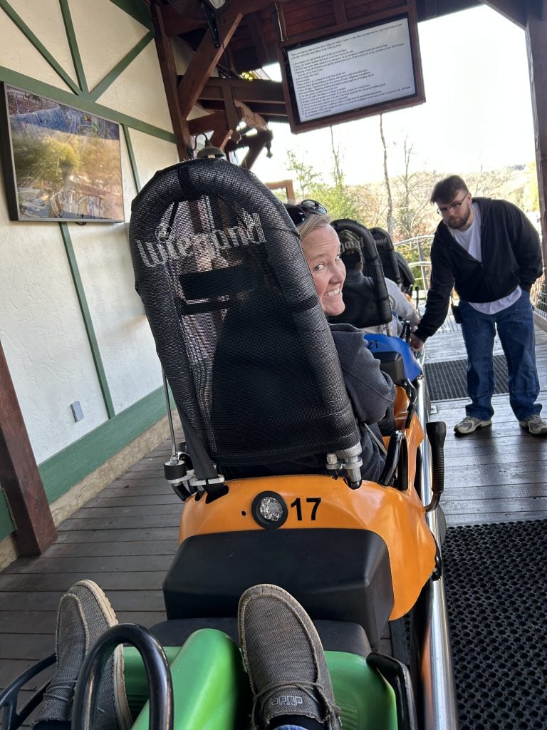 Close-up view from a rider’s perspective on the Georgia Mountain Coaster in Helen, GA, showing a row of alpine coaster cars lined up at the loading station. A smiling woman in car number 17 looks back, while a staff member assists other riders ahead under a chalet-style canopy.