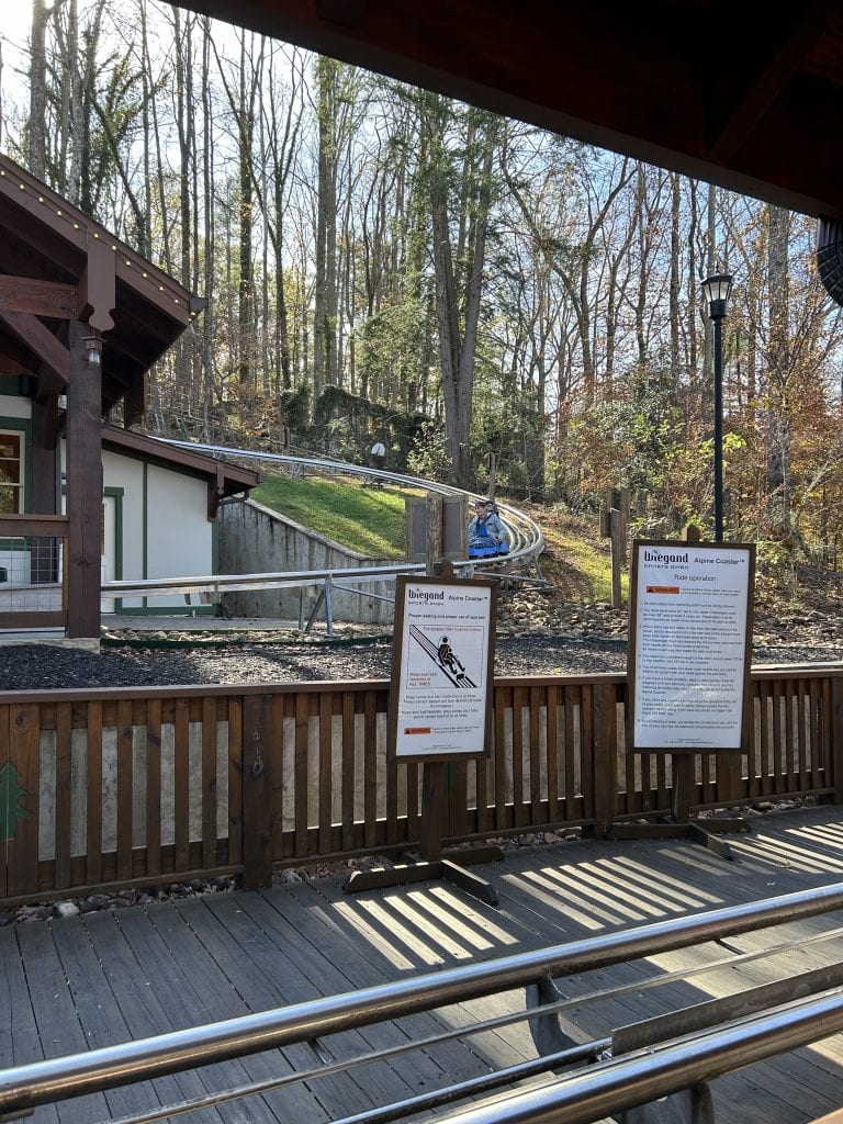 View of the Georgia Mountain Coaster track curving through a wooded hillside near the loading station in Helen, GA, with alpine-style buildings and safety signage in the foreground. A rider in a blue cart is approaching the platform as sunlight filters through the bare winter trees.