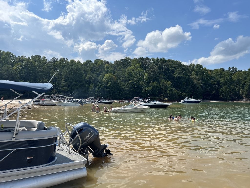 A lively summer scene on Lake Lanier shows families swimming and wading in shallow water while boats anchor along the sandy shoreline backed by dense forest. The sky is bright with scattered clouds, and a pontoon boat with a Yamaha motor is docked in the foreground.