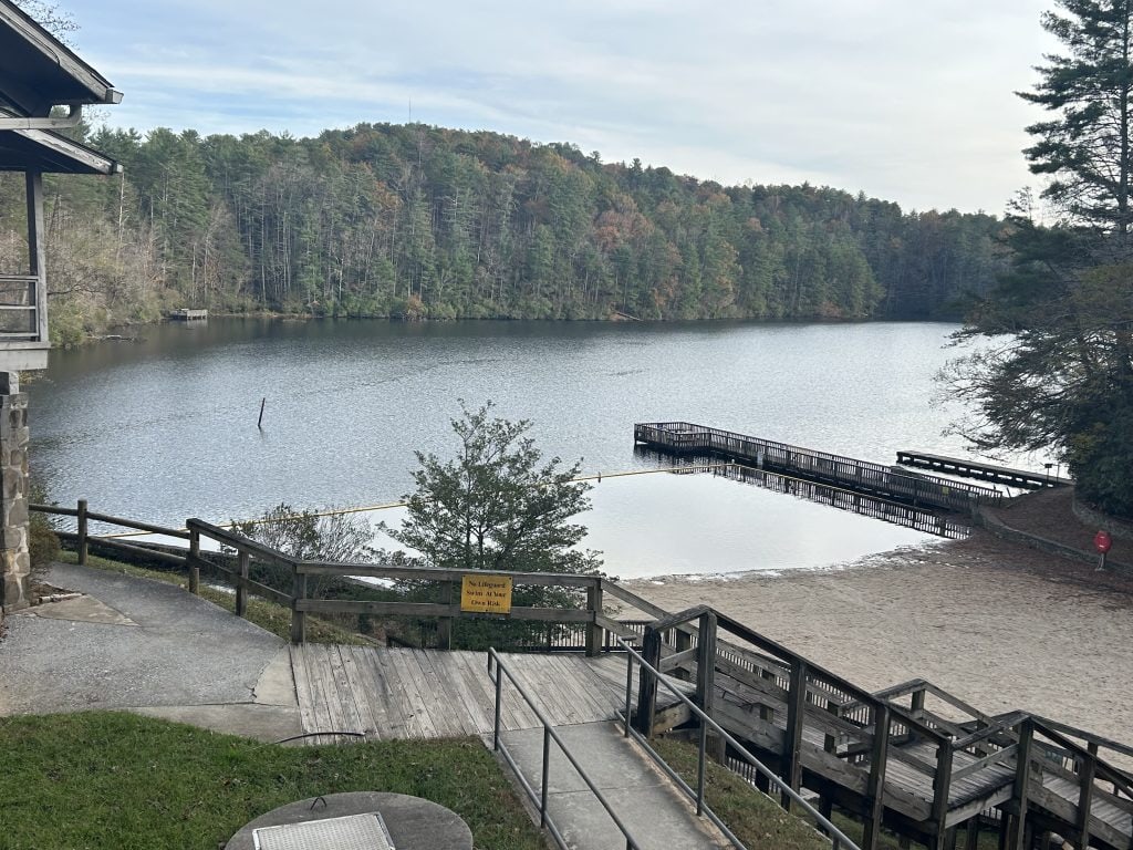 Peaceful view of the lake at Unicoi State Park in North Georgia, featuring a sandy beach area, wooden boardwalks, and a long fishing or swimming dock extending into the calm water. The lake is surrounded by dense forest with trees showing hints of fall color under an overcast sky.