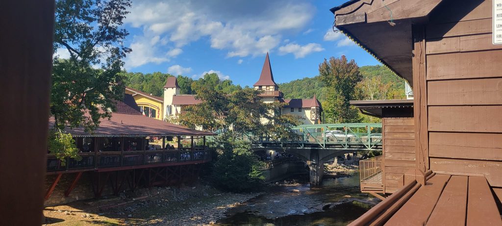 Scenic view of downtown Helen, Georgia, featuring alpine-style buildings with steep red roofs and pointed towers nestled along a gently flowing river. A green pedestrian bridge spans the creek, connecting charming shops and restaurants surrounded by lush trees and blue skies.
