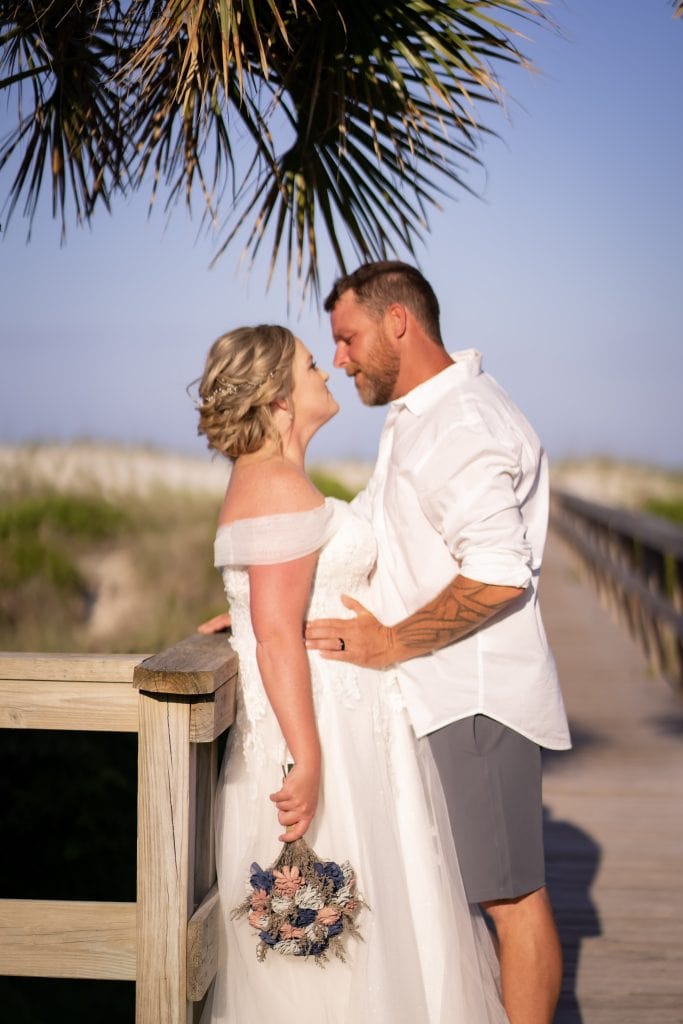 Bride and groom share an intimate moment on a sunlit wooden boardwalk with sea oats and dunes in the background. The bride holds a bouquet of dried flowers in soft pink and blue tones while the groom, dressed in a casual white shirt and shorts, embraces her under a palm frond.