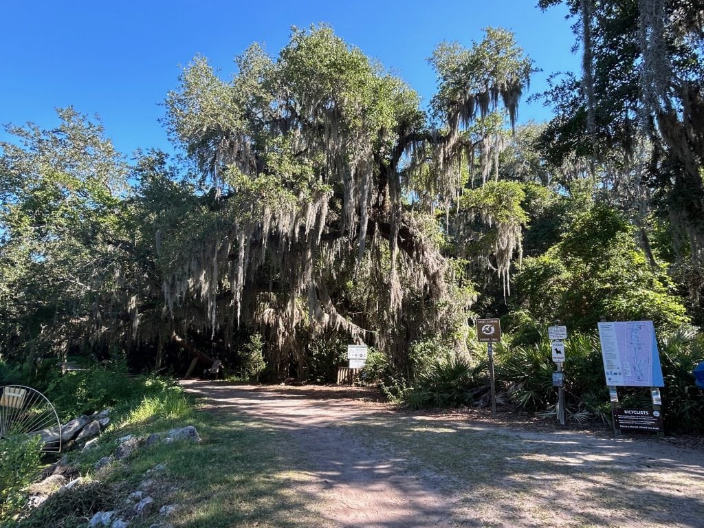 Entrance to Egan's Creek Greenway on Amelia Island, with a dirt trail leading into dense trees draped in Spanish moss. Informational signs and a large trail map are posted near the shaded path under a clear blue sky.
