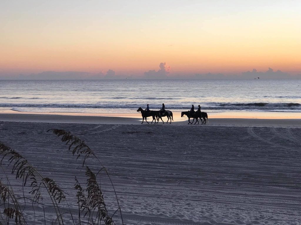Four horseback riders silhouetted against a pastel sunrise on a quiet beach, with gentle waves and smooth sand in the foreground. This peaceful coastal scene captures the natural beauty of Amelia Island.