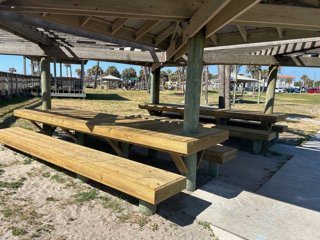 Covered wooden picnic pavilion with built-in benches and tables set on sandy ground near a grassy park area. Palm trees, a playground, and nearby parking are visible in the background under a sunny blue sky.