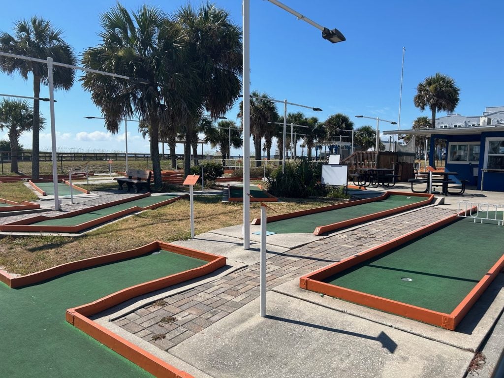 Outdoor mini golf course with green turf and orange borders, surrounded by palm trees and picnic tables near a blue concession stand. Located by the beach on Amelia Island, offering a fun activity with ocean views.