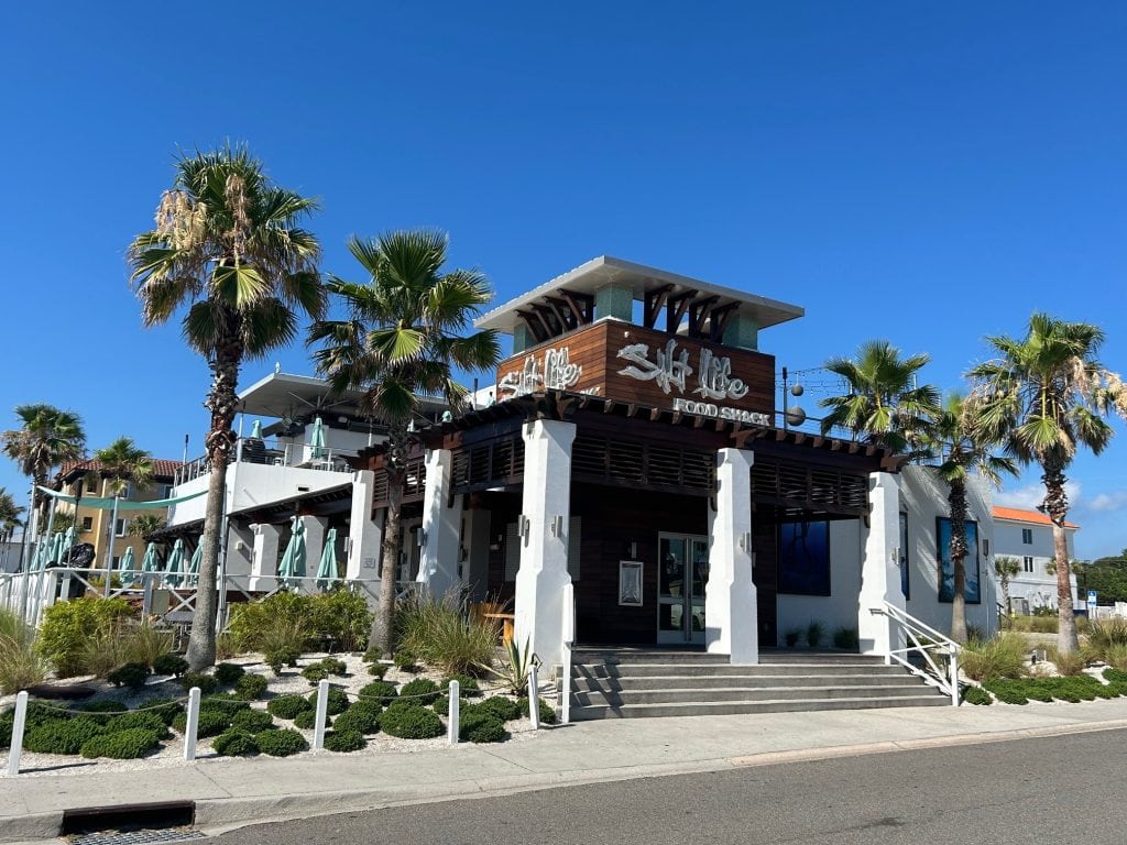 Exterior of Salt Life Food Shack on Amelia Island, a modern coastal restaurant with white stucco walls, wood accents, and a rooftop dining area. Surrounded by palm trees and beach-style landscaping, the building sits under a clear blue sky.
