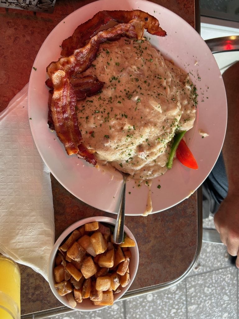 Hearty breakfast plate from Tortuga Jack’s in Amelia Island featuring crispy bacon, creamy sausage gravy over biscuits, and a side of golden brown home fries. Garnished with chopped herbs and a slice of tomato and green pepper for color.