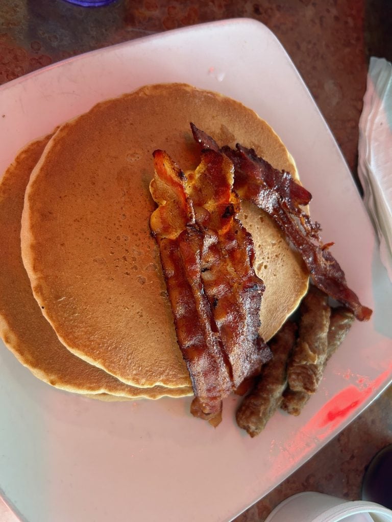 Breakfast plate from Tortuga Jack’s in Amelia Island featuring two golden pancakes topped with crispy bacon strips, served with a side of savory sausage links. The meal is presented on a white square plate with a soft, warm lighting ambiance.