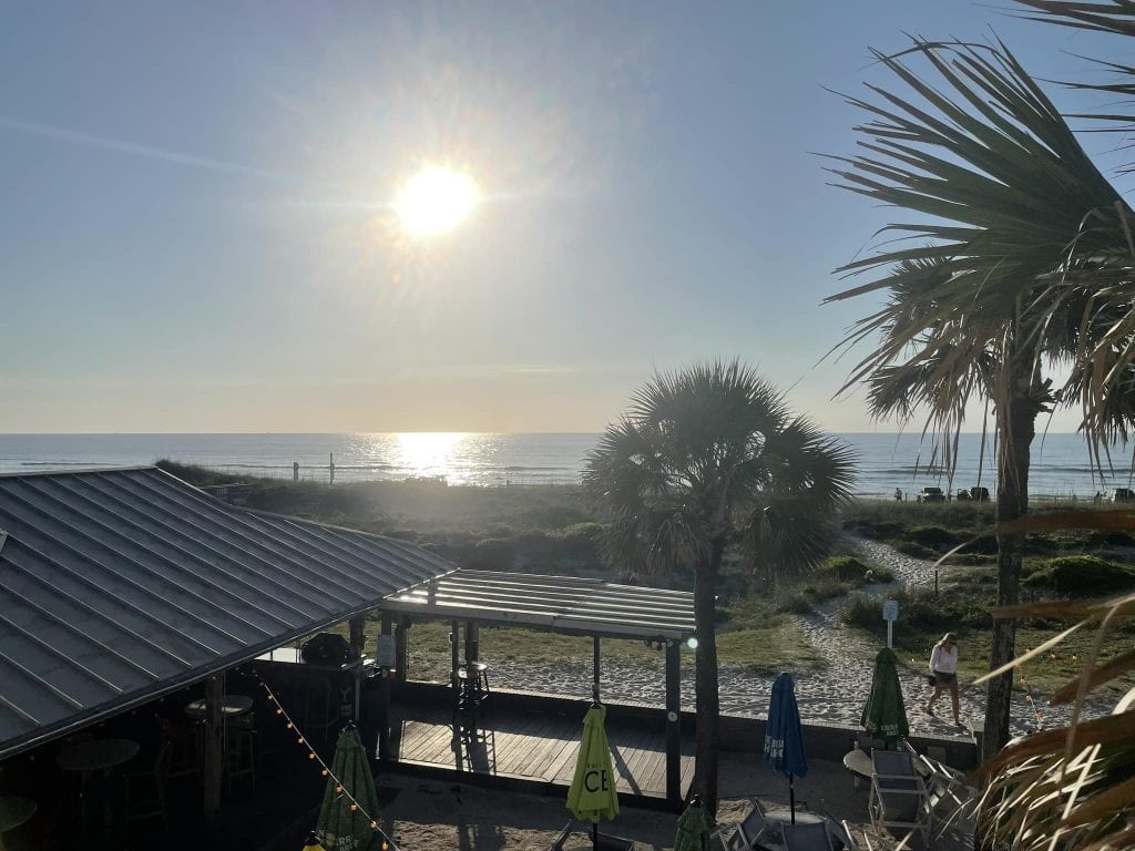 View of the beach on Amelia Island during golden hour with the sun low in the sky reflecting off the ocean. A beachside bar with umbrellas and string lights sits in the foreground, while palm trees and a sandy path lead to the shoreline.
