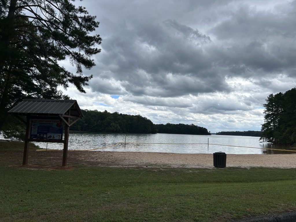 A sandy swimming beach at Bald Ridge Campground faces a calm lake under a cloudy sky, with forested shorelines in the distance. A covered sign near the water provides life jacket safety information, and a trash can sits near the grassy edge.
