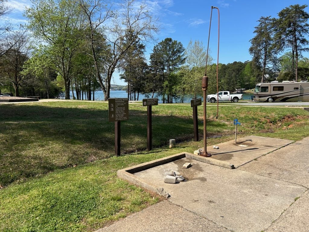 A dump station at Bald Ridge Campground features a concrete pad with sewer and water hookups, surrounded by grassy areas and signage with usage instructions. In the background, RVs are parked near the lake under a clear blue sky with trees lining the shoreline.