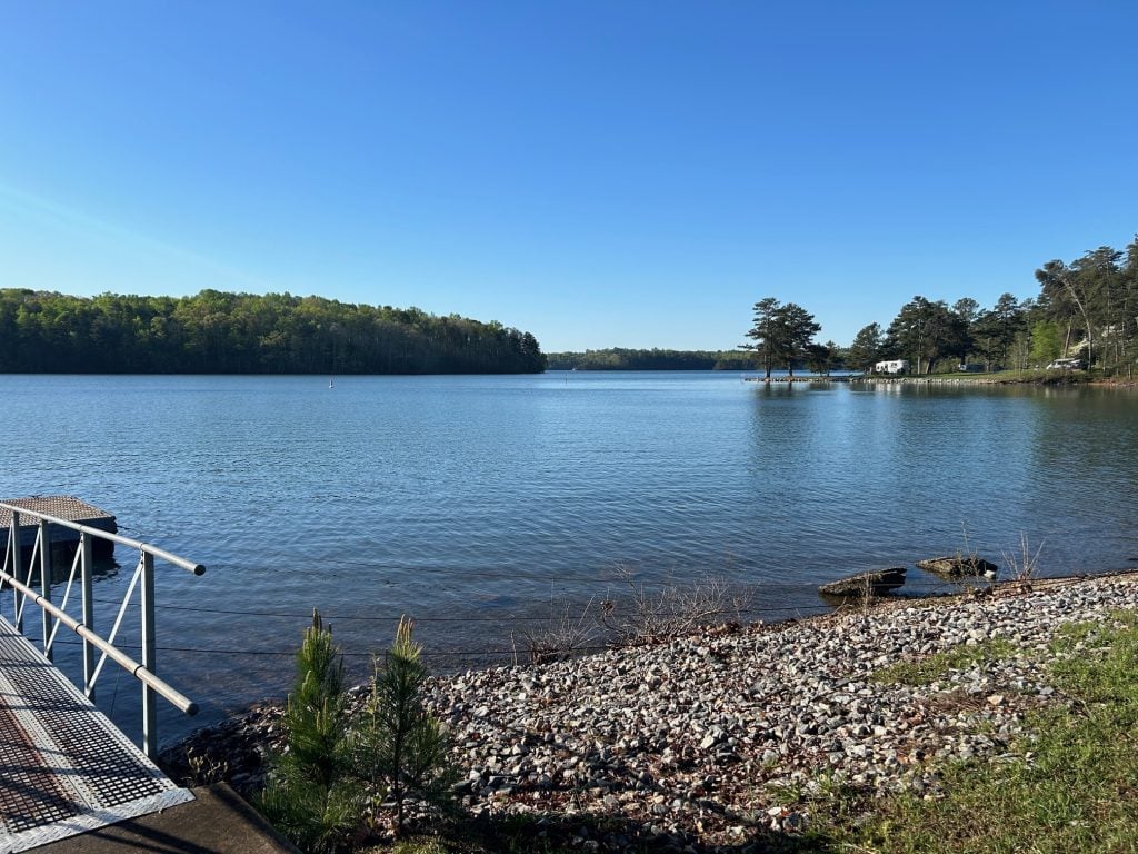 A peaceful lakeside view at Bald Ridge Campground shows calm blue water bordered by a rocky shoreline and a metal dock on the left. In the distance, pine trees line the shore with a few RVs and a small pier visible under a clear blue sky.