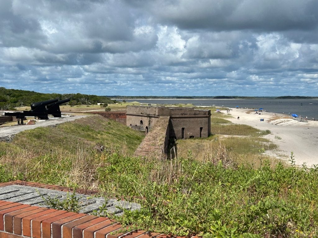 View of Fort Clinch on Amelia Island featuring historic brick fortifications, grassy embankments, and black cannons facing the shoreline. The scene extends to a sandy beach with scattered beachgoers and umbrellas under a cloudy sky.