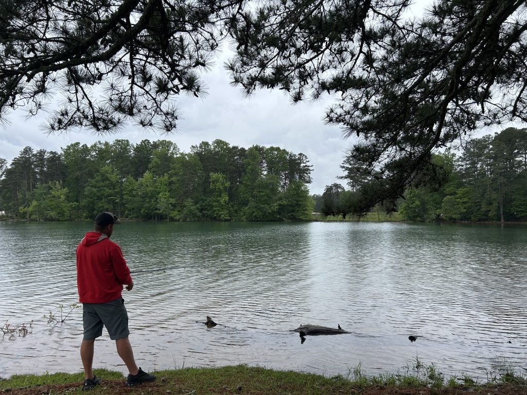 A man in a red hoodie stands at the edge of the water fishing at Bald Ridge Campground, surrounded by pine trees and calm lake water. Overhanging branches frame the scene, with dense green forest lining the opposite shore.
