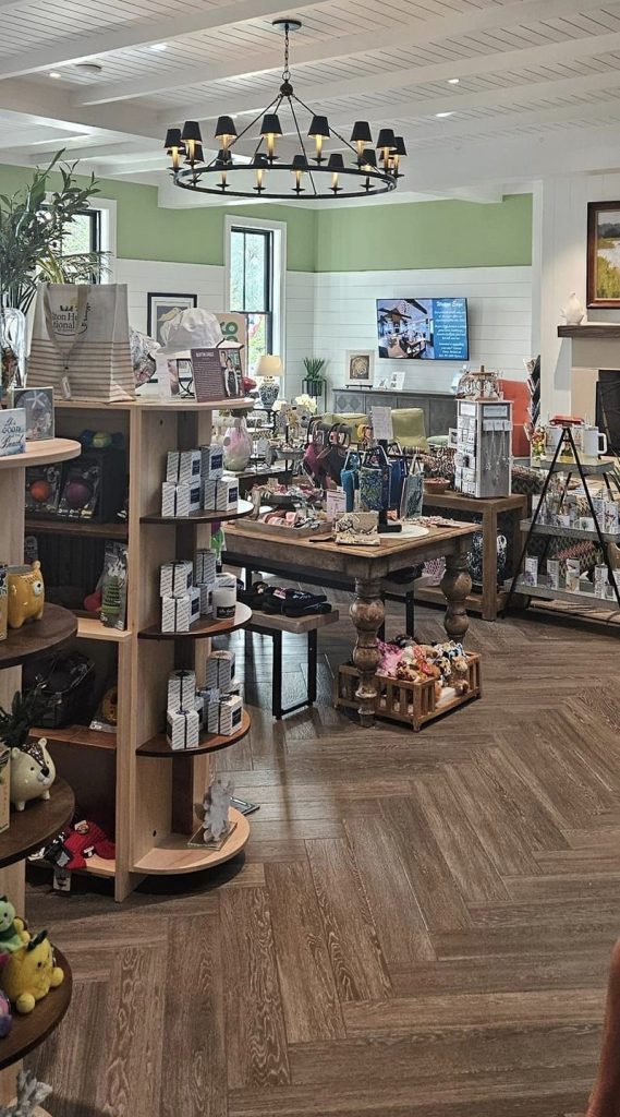 Gift shop at Hilton Head National RV Resort featuring wooden shelves and tables filled with souvenirs, toys, candles, and bags. The space has a modern coastal design with wood-look flooring, green and white walls, and a black chandelier hanging from a white plank ceiling.