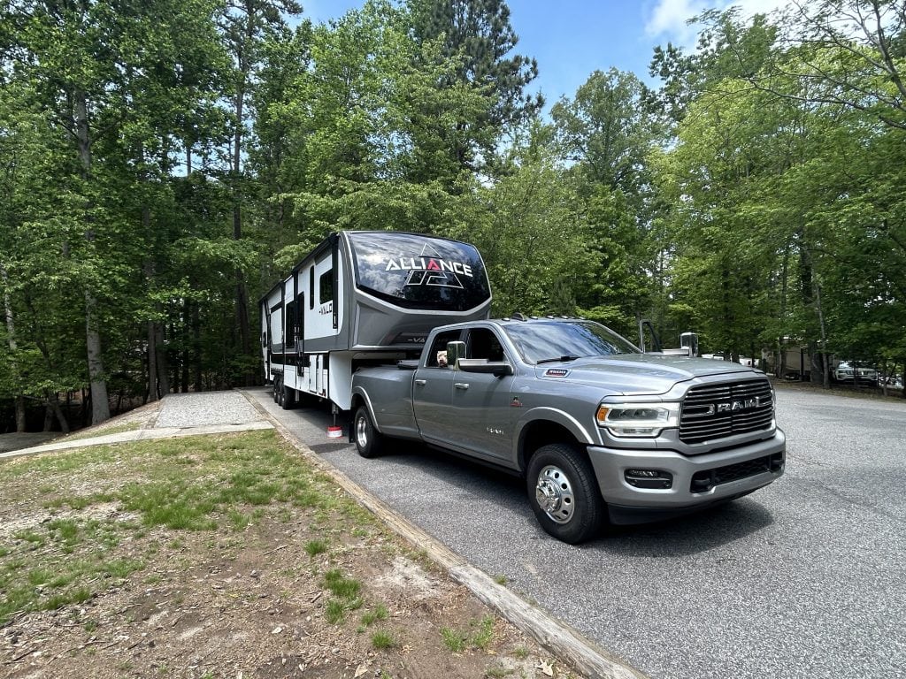 Back-in RV site at Shady Grove Campground with a large Alliance Valor fifth-wheel trailer hitched to a silver RAM dually truck. The gravel pad is surrounded by trees and set beside a paved loop.