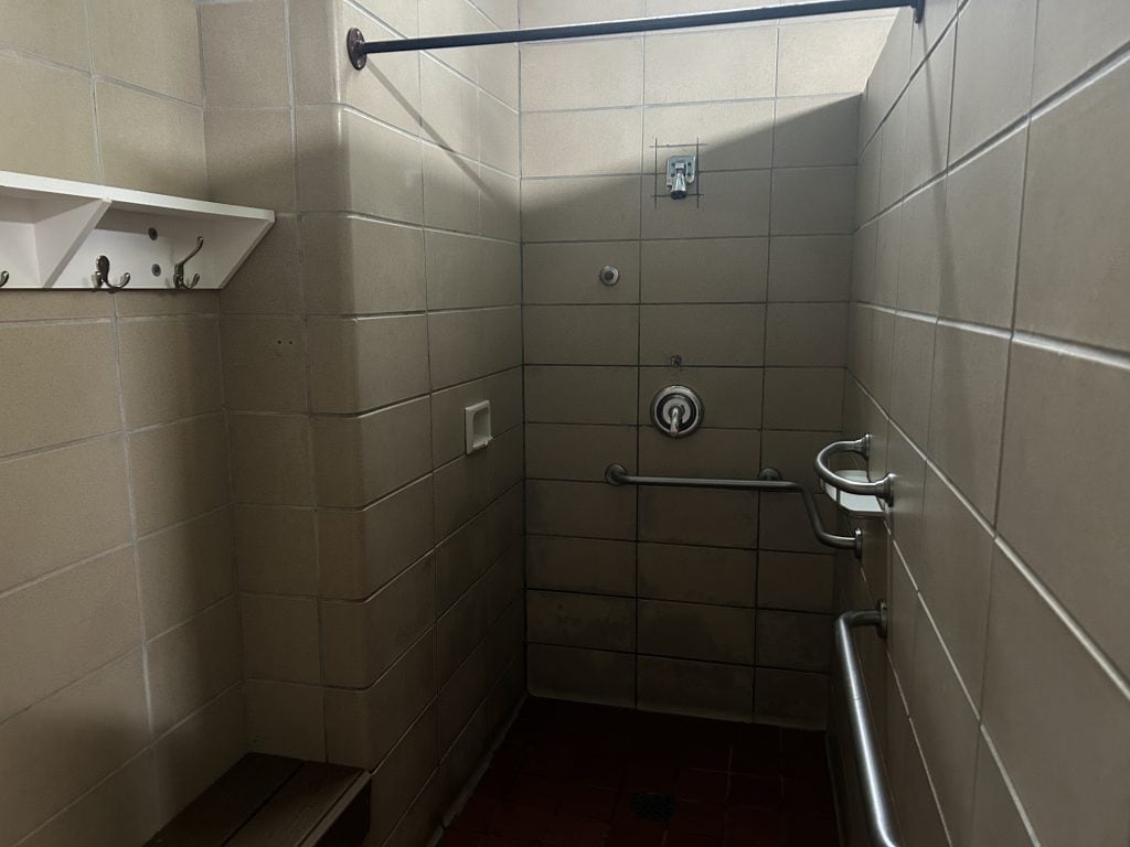 Accessible shower stall at Shady Grove Campground with tiled walls, grab bars, a bench, wall hooks, and a showerhead above a drain in the red tile floor. A curtain rod is installed overhead for privacy.