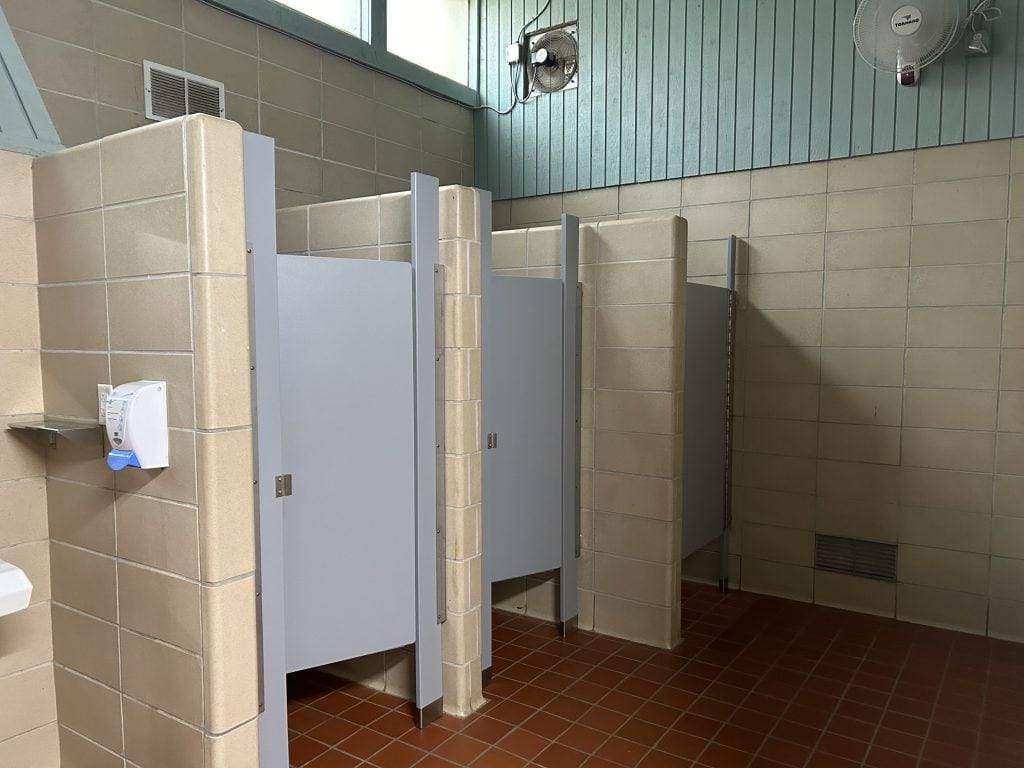 Clean but dated restroom at Shady Grove Campground featuring three toilet stalls with light gray doors, tan tiled walls, and red tile flooring. A soap dispenser and wall-mounted fans are visible, along with natural light coming from high windows.