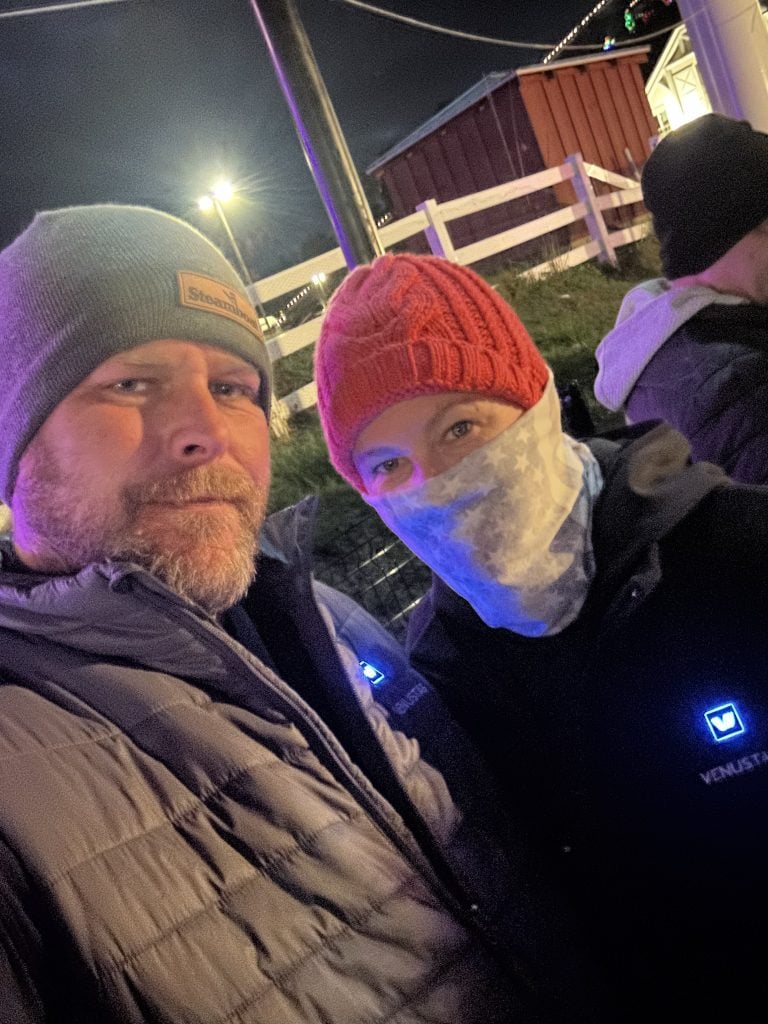 Man and woman bundled up at night, both wearing Venustas heated vests with glowing blue power indicators. They are dressed warmly with hats and face coverings, standing near a white fence and red barn under festive outdoor lighting.