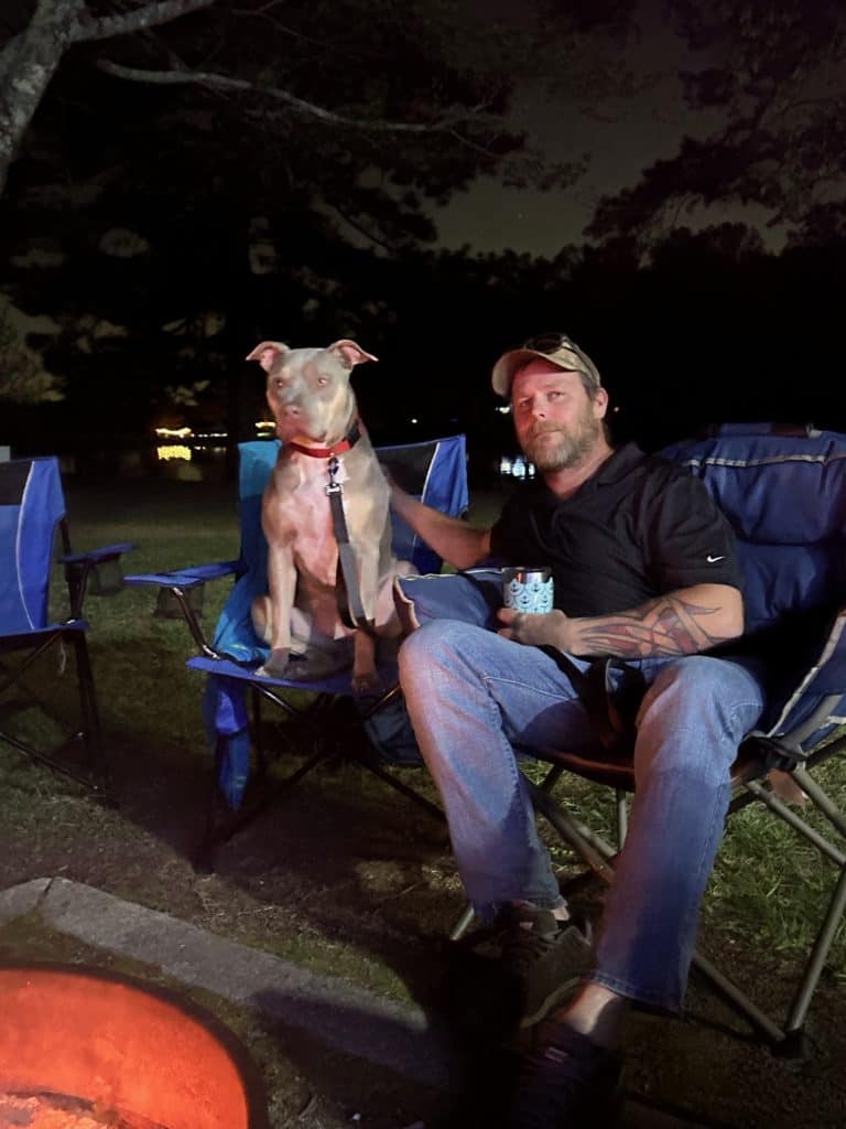 A man sits in a blue camping chair at night beside a fire pit, holding a mug and resting his hand on a gray dog with a red collar seated in the chair next to him. The warm glow of the fire lights their faces, with dark trees and faint reflections on water in the background.