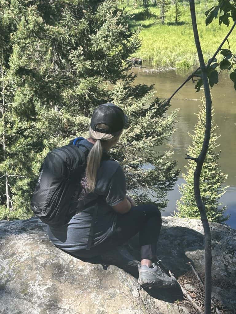 A woman wearing a black cap, gray shirt, black leggings, and hiking shoes sits on a rock ledge with a black backpack. She looks out over a pond partially hidden by evergreen trees, where a moose stands in the shallow water near the grassy shore.