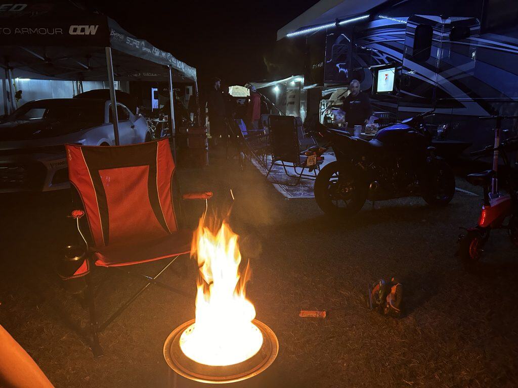 A small campfire burns in a Solo Stove in the foreground beside a red folding chair at a nighttime campsite. Behind it, a motorcycle, a large RV with blue accent lighting, and a car under a canopy are visible, with people gathered under the awning.