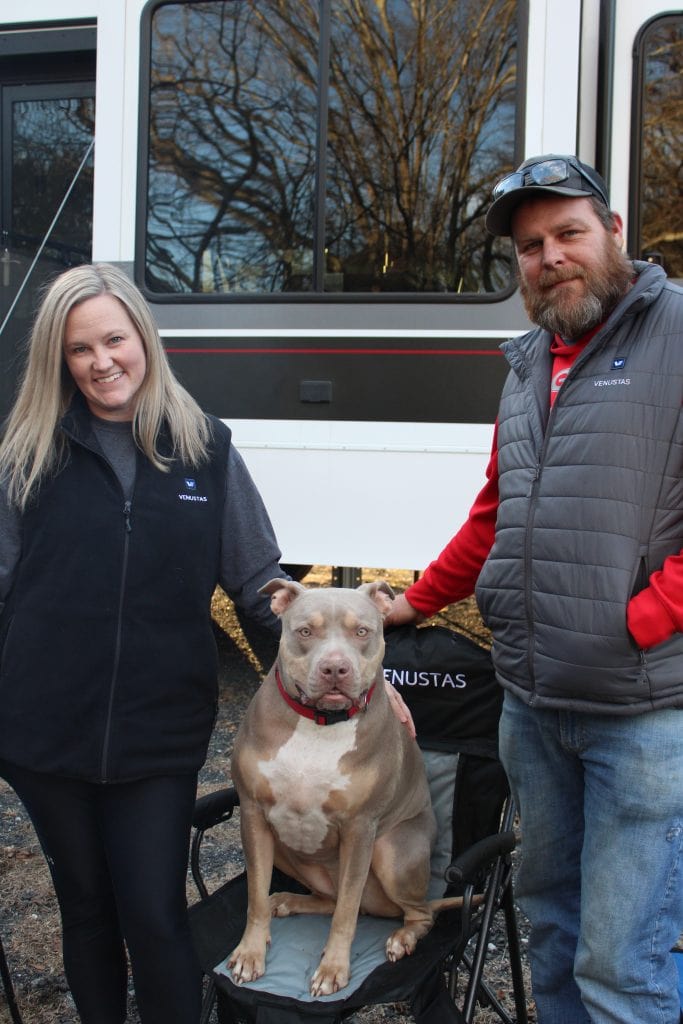 Morgan and Alan stand on either side of their dog, Coco, who’s sitting proudly on a camp chair in front of their RV, with bare winter trees reflected in the window behind them.